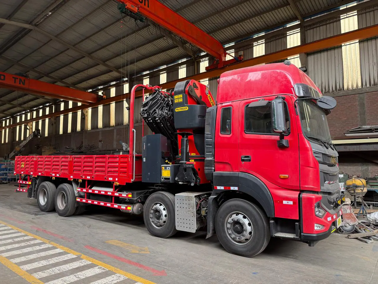Red JAC 8x4 knuckle-boom crane truck viewed from the front three-quarter inside a fabrication workshop